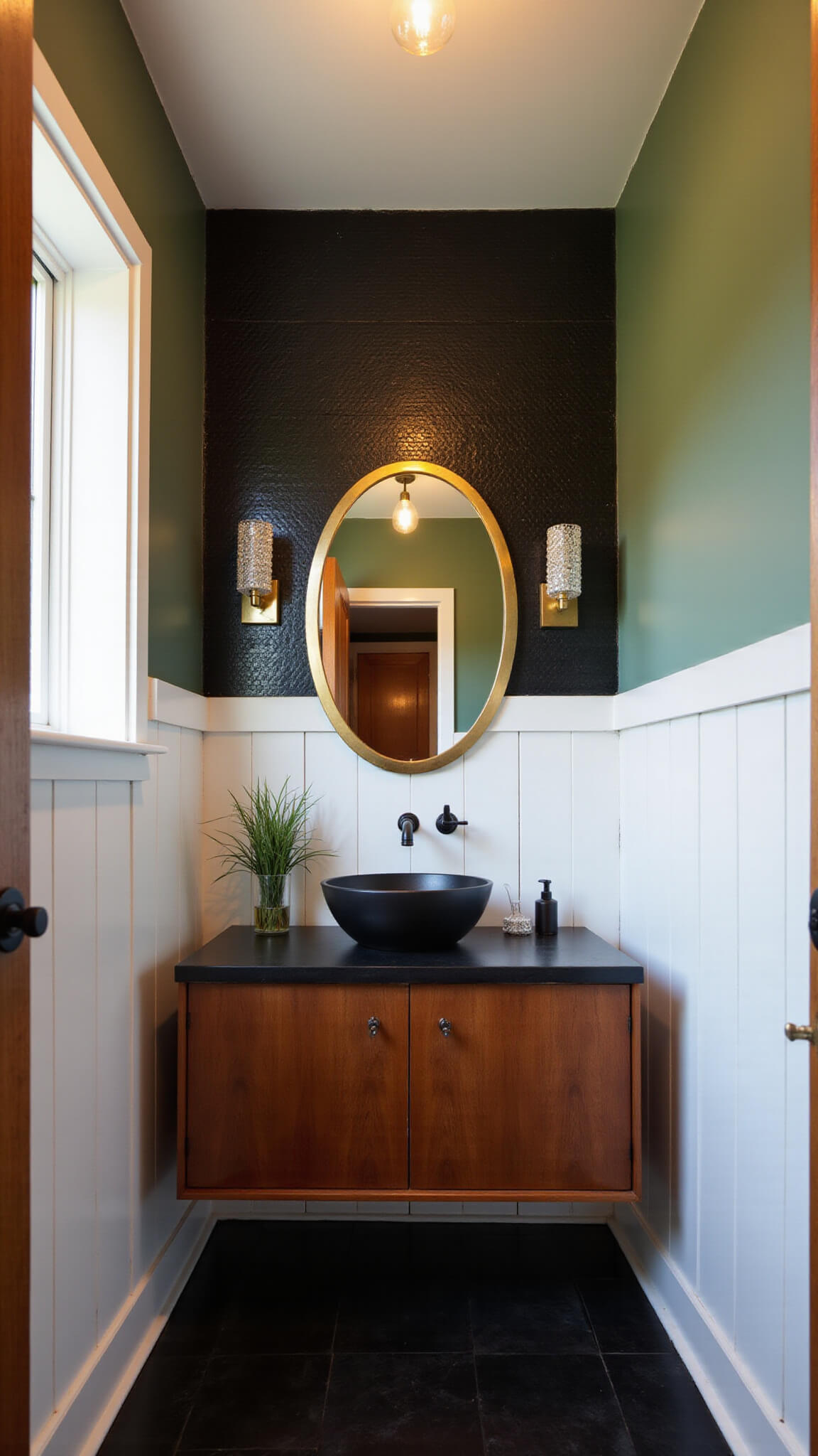 Low-angle view of a moody powder room with black penny tile, sage green walls, floating teak vanity, matte black vessel sink, brass mirror, crystal sconces, and Edison bulb chandelier.