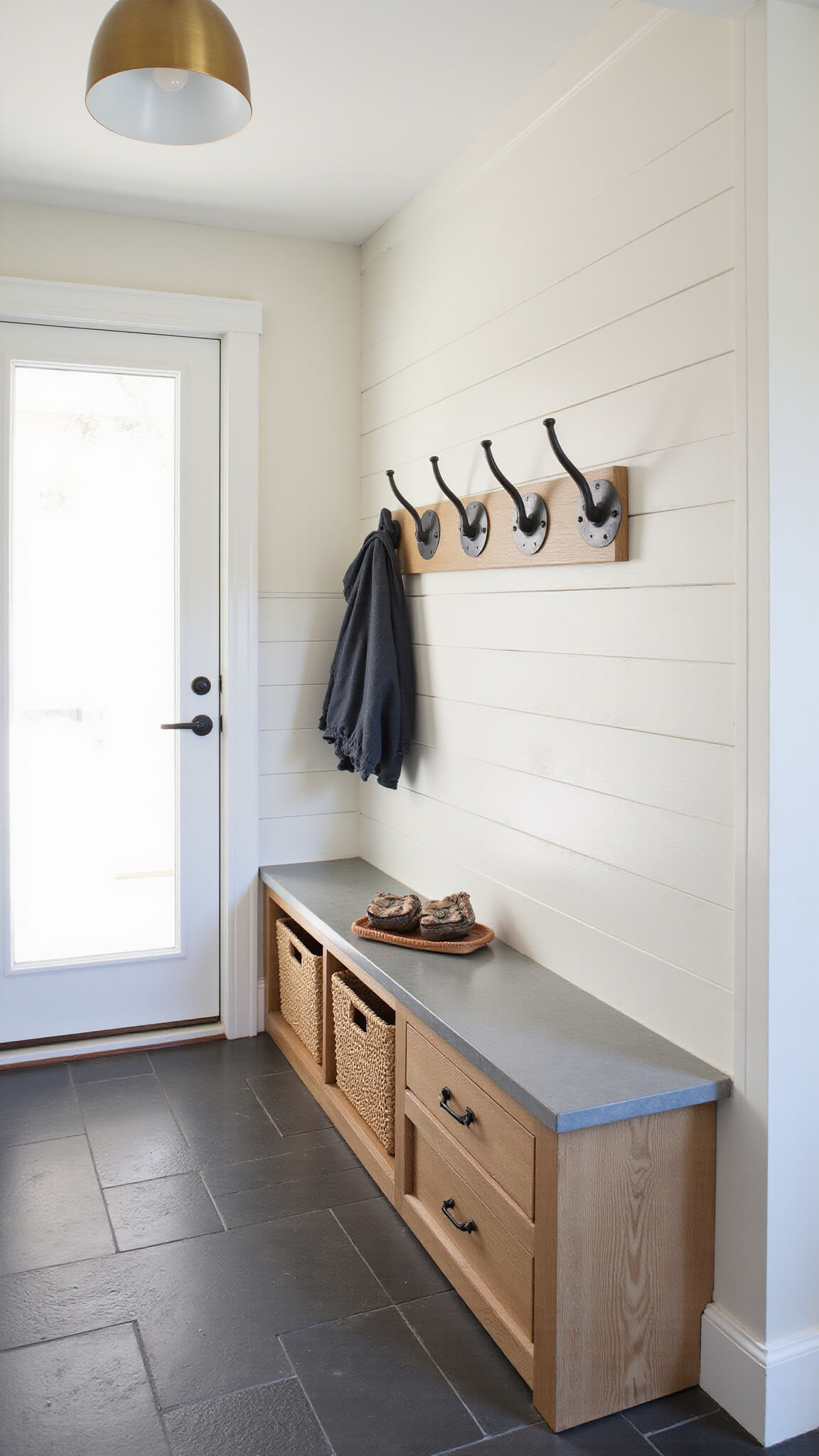 Entryway mudroom with custom steel and oak hooks on shiplap wall, built-in bench with drawers, woven baskets, slate floor, and brass pendant light.
