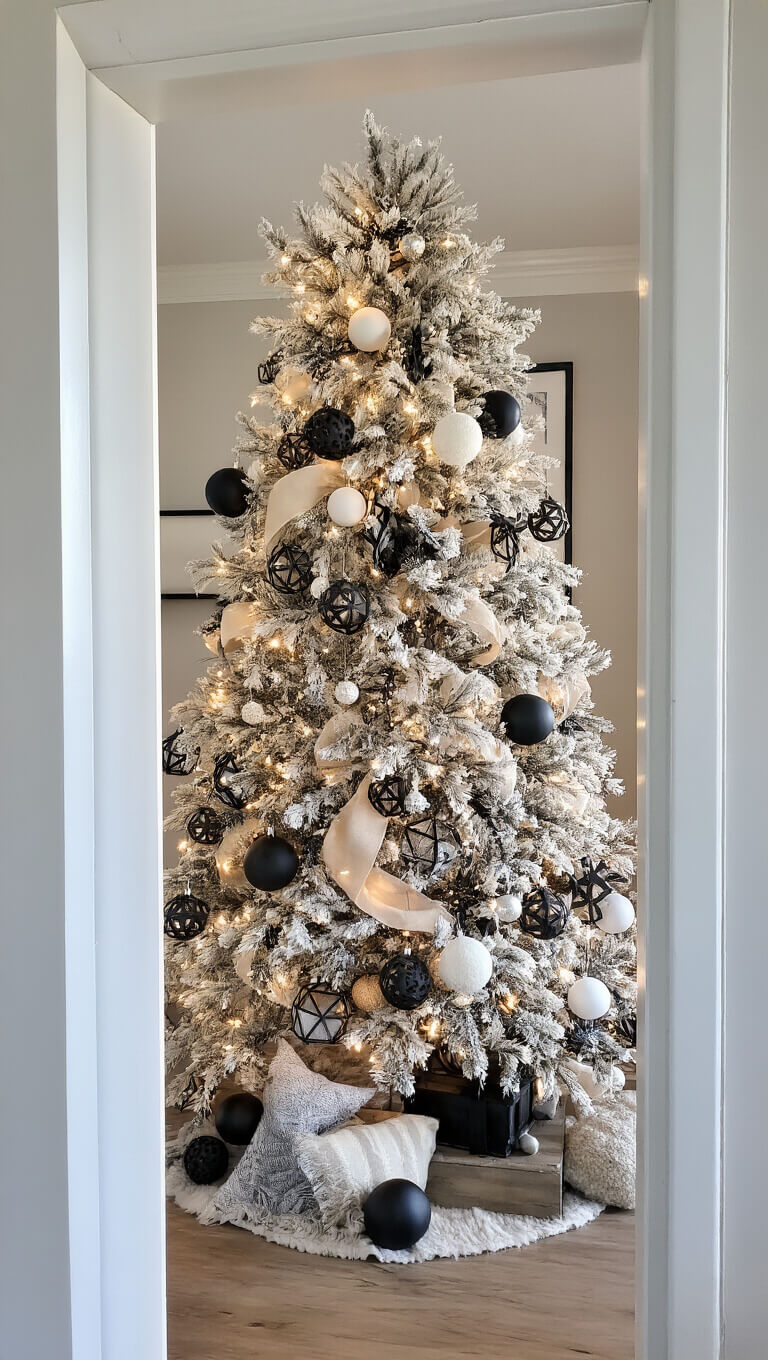 Modern farmhouse living room with 8.5ft flocked Christmas tree decorated in black, white, and champagne matte ornaments, geometric metal accents, and soft fabric pieces; viewed through doorway with warm late afternoon light and spotlights.