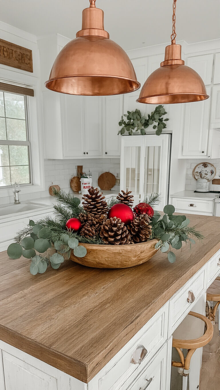 Overhead view of a 6x8ft kitchen island with a distressed wood top and white base, styled with a holiday display in a wooden dough bowl filled with pinecones, red ornaments, and eucalyptus, under copper pendant lights in warm morning light.