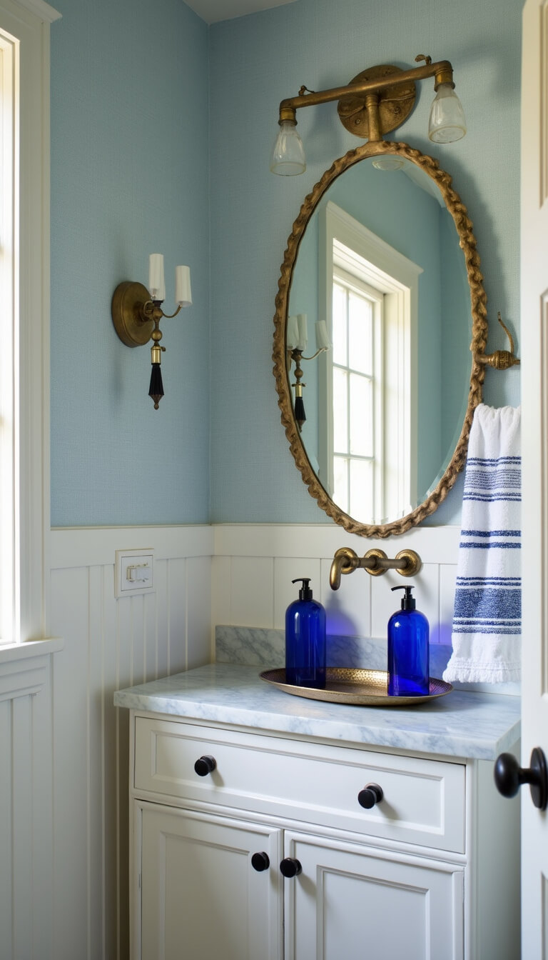 Small powder room with pale blue grasscloth wallpaper, white wainscoting, marble vanity, vintage brass mirror, cobalt blue soap dispensers, hammered metal tray, and striped Turkish hand towels, softly lit from corner angle.